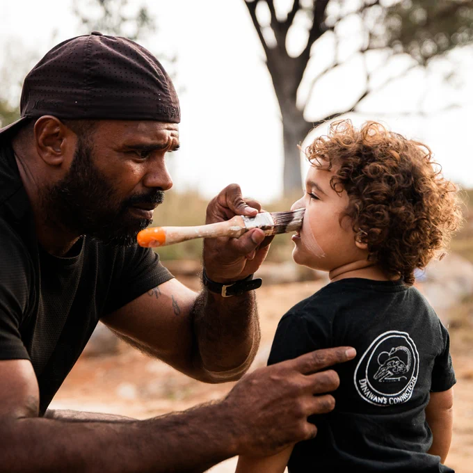 Man painting ceremonial paint on young girls face in front of a tree