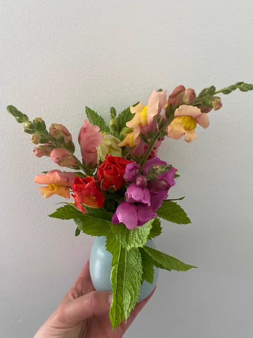 A small teal vase held by a hand. Bouquet of mint, orange roses, and snapdragons of various colors. 