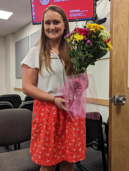 A girl (Ali Cala) is standing up and smiling towards the camera while holding a flower bouquet composed of multiple different flowers with yellow, red, pink, and white colors. She has long straight light brown hair and is wearing a white top with a red-orange color skirt with white dots. 