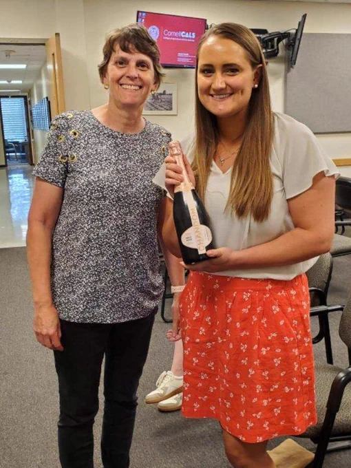 Two women (Chris Smart on left and Ali Cala on right) are both standing up and smiling towards the camera. A woman on the left has short dark brown hair and is wearing a gray & black top with black jeans. A woman on the right has long light brown hair with white top and orange-red skirt with white dots and is holding a bottle. The background looks like an indoor conference room.