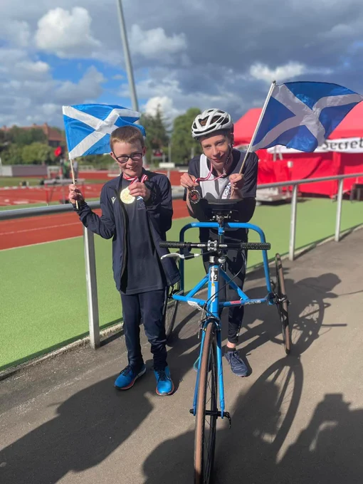 Photo of Ian and Matthew smiling with one of their medals each. They also have a mini Scottish flag on a stick each. Ian is standing on left, while Matthew is on his blue frame runner in the centre. The track is in the background