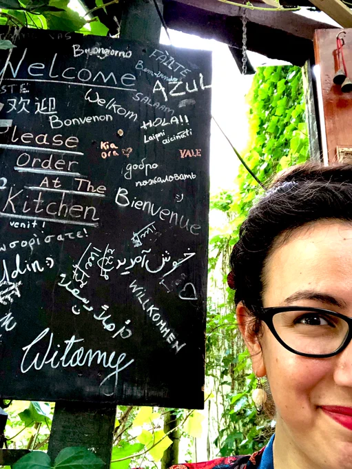 Partial selfie: half of my face next to a blackboard with “Welcome” written on it in several different languages, against a background of illuminated greenery.