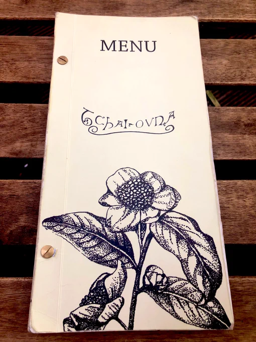 A closed Tchai Ovna menu on a slatted wooden table. The menu features a stylized flower with two blossoms and three leaves growing from a central stem.