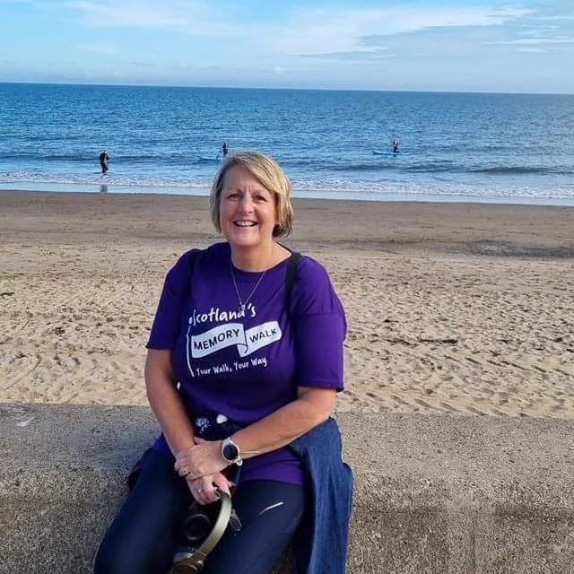 Susan sits on the wall at the beach and smiles at the camera. She is wearing a purple Memory Walk t-shirt. 