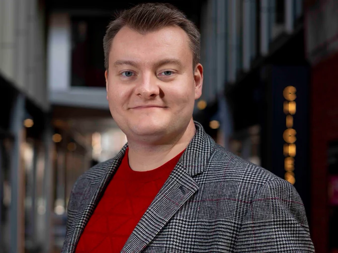 Matthew dressed in a black and white checkered blazer and a red top. Smiling at the camera.