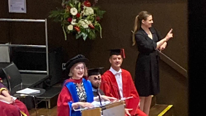 Woman in academic robes delivers speech from lectern. Interpreter, standing, signs next to her. 