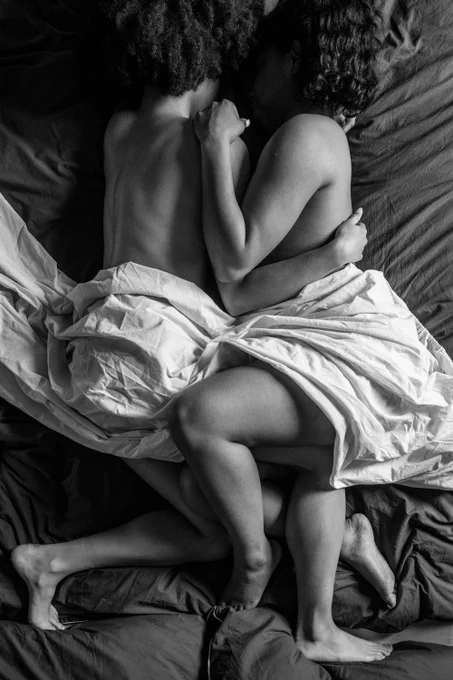Two women lie in a bed together with their legs tangled and sheets around their waist, their faces are obscured. 