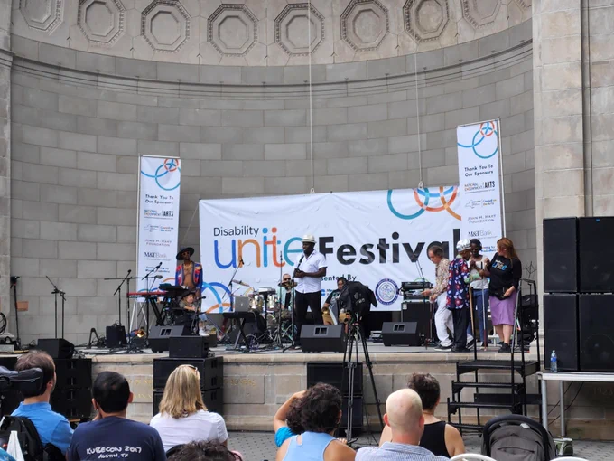 A group of people standing on a stage with a Disability Unite background and musical instruments, looking at a crowd.