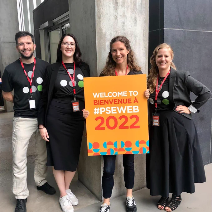 Four people holding up an orange sign that says "Welcome to/Bienvenue a #PSEWEB 2022"