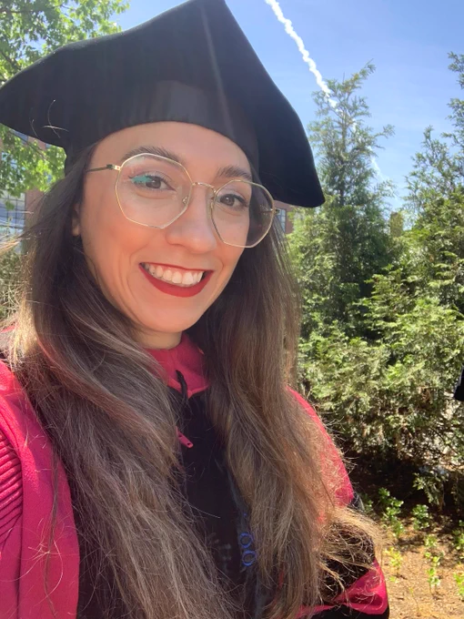 A photograph of Vanessa Sanchez, a brown haired woman with gold glasses smiling, at the camera. She is wearing a red graduation gown with black graduation cap.