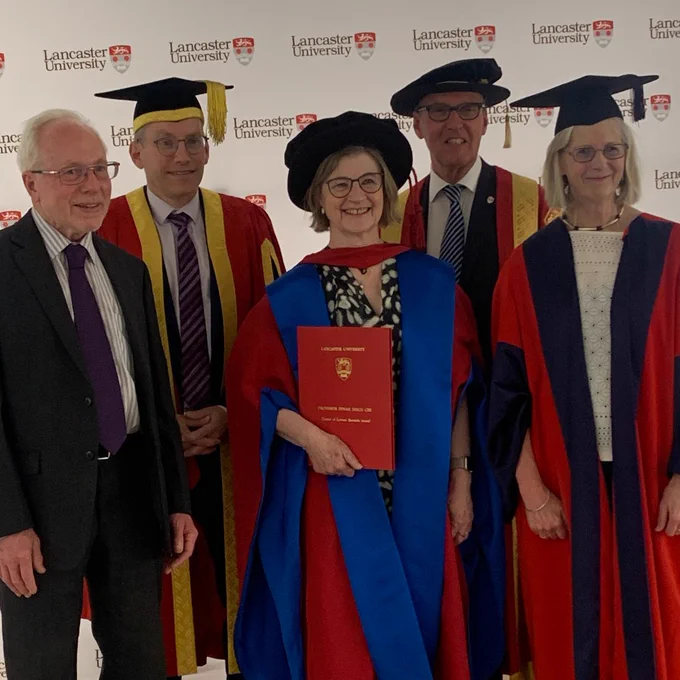 Five people standing in a line in formal dress, with four wearing academic robes, two with mortarboards and two with PhD caps. Central figure holding honorary degree certificate. 
