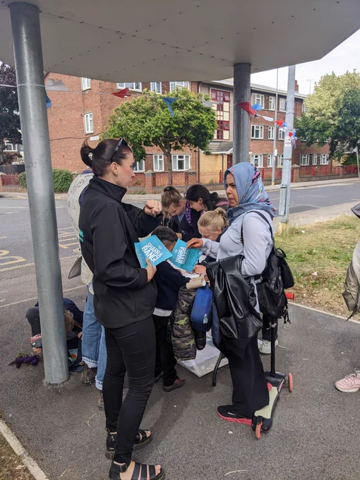 A group of people chatting, with some children drawing on a piece of paper on a table. 