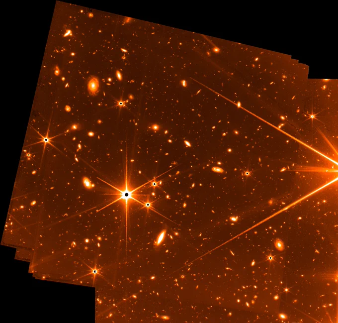 Bright stars (each with 6 spikes of light radiating from them) dominate the foreground, while the background is littered with galaxies.  The image is reddish in tone, selected to optimize visual contrast. Mission managers have also been using this during Webb's commissioning to differentiate engineering images from the science images still to come.