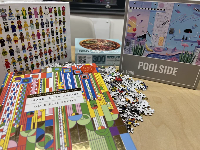 3 jigsaw sets on a table: 1 of them has a picture of a bunch of lego mini figures, the second one has a 90s aesthetic poolside scene and the third one an abstract colourful grid design.