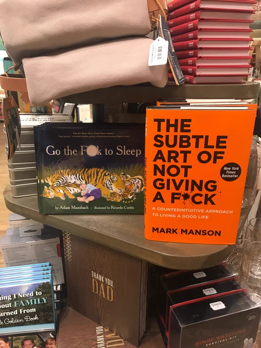 A bookshelf in a bookstore. On the featured shelf, two books: GO THE F🌙CK TO SLEEP and THE SUBTLE ART OF NOT GIVING A F*CK