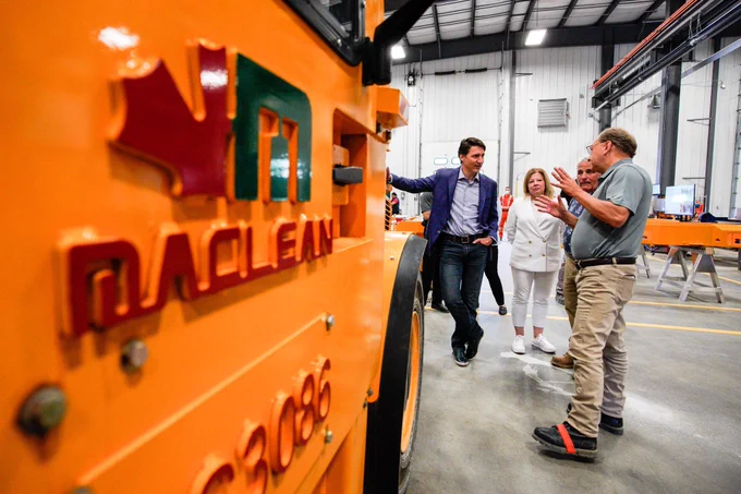 Prime Minister Justin Trudeau is standing beside a large industrial vehicle. On his left side, Members of Parliament Viviane Lapointe and Marc Serré are standing. An employee is standing beside them and gesturing with his hands.