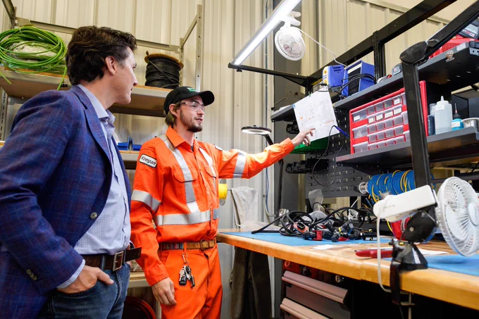 Prime Minister Justin Trudeau is standing with his hands in his pockets. On his left side, an employee is pointing at a piece of paper clipped onto a shelf.
