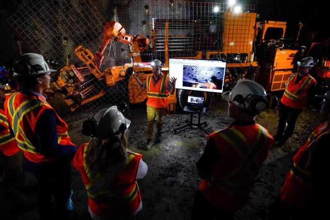 Prime Minister Justin Trudeau is standing in a semi-circle with other people. They are facing an employee, who is talking and gesturing with their hands, and a television screen that is showing a presentation. Behind the screen, a large industrial vehicle is visible.