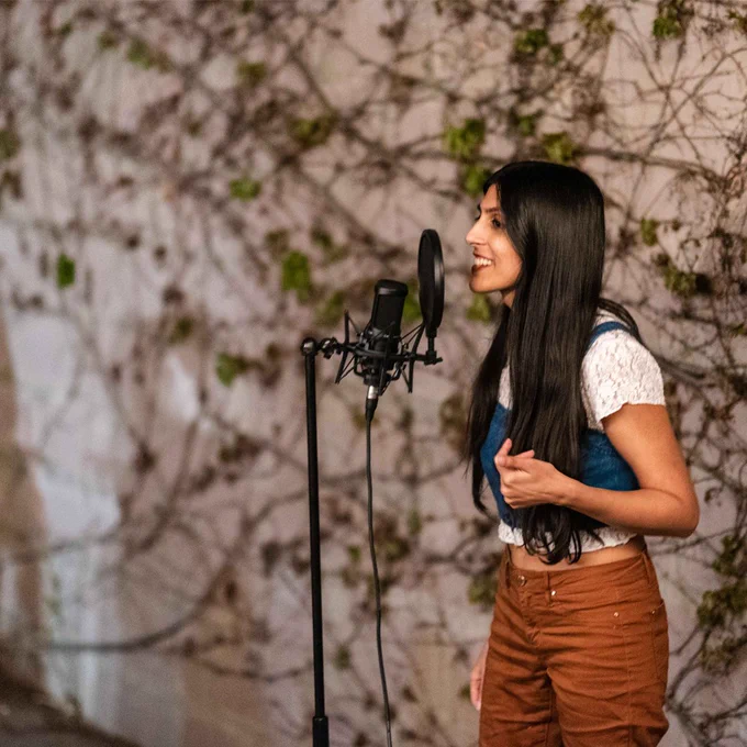 Zen is standing next to a mic, gesturing with one hand. Behind her a wall is covered in vines. 
