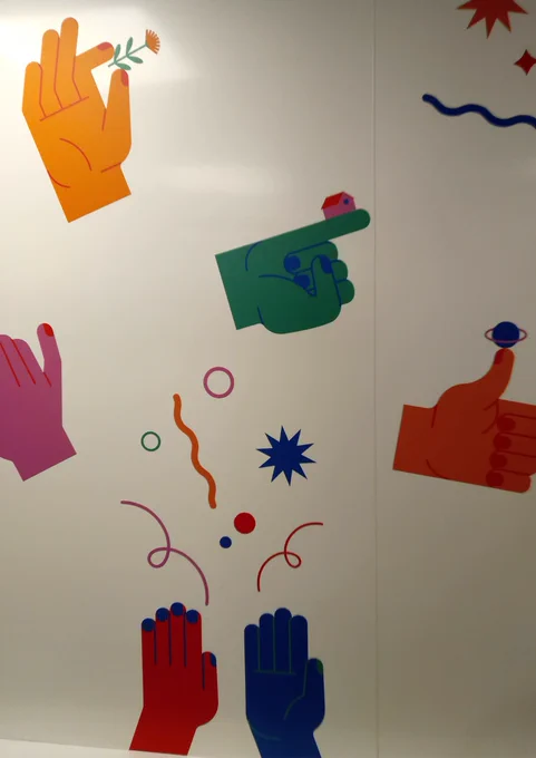 On a white bathroom wall, there are vinyls of hands in different colours - from orange to red, pink, green and blue. One hand balances a little house, another one a planet and another one holds a flower.