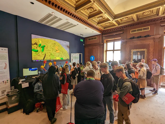 A picture of a crowd visiting the microbial Puppet Masters exhibit at the royal society. There is a screen showing a video of bacteria and viruses and 3d printed models on show.