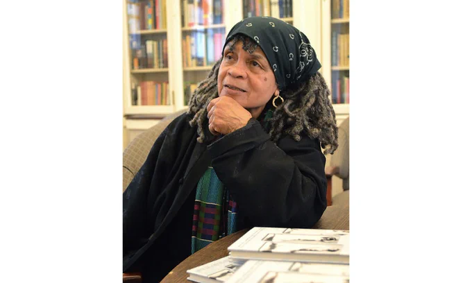 Poet Sonia Sanchez sits in a chair in a well lit library. She rests her elbow on a table upon which three copies of her book are displayed. 