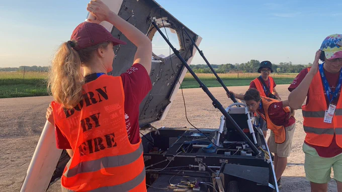 Students in safety vests point the MIT solar car at the sun with a green field in the background
