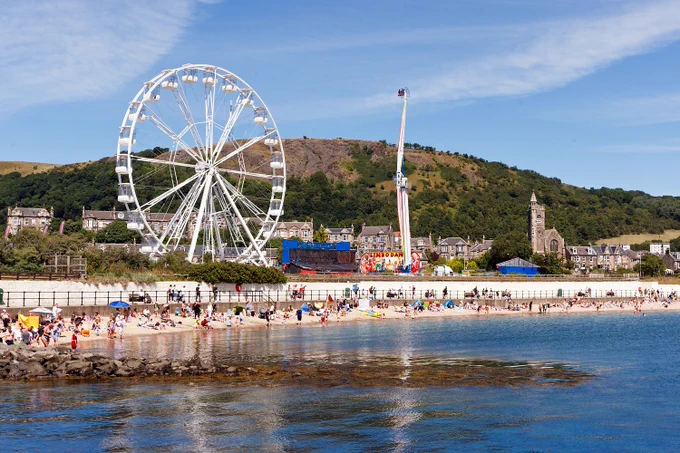 High tide at Burntisland beach, busy with people, the fairground's big wheel in the background