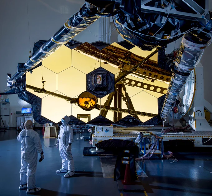 The Webb telescope's primary mirror is inside a facility laying on its side, with engineers in white protective suits. The mirror's two wings that give it its honeycomb shape are not deployed in this image. Its secondary mirror, held by a long boom, is deployed in front of the primary mirror. The primary mirror is made up of multiple hexagonal mirrors assembled into one shape, and are covered in gold. 