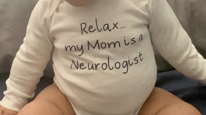 Baby in a white onesie that says “Relax… my Mom is a Neurologist”