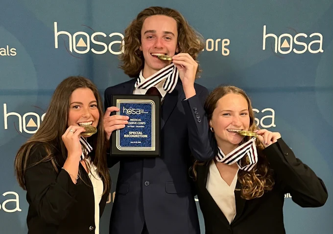 Three students biting into medals and holding an award plaque in front of HOSA logo