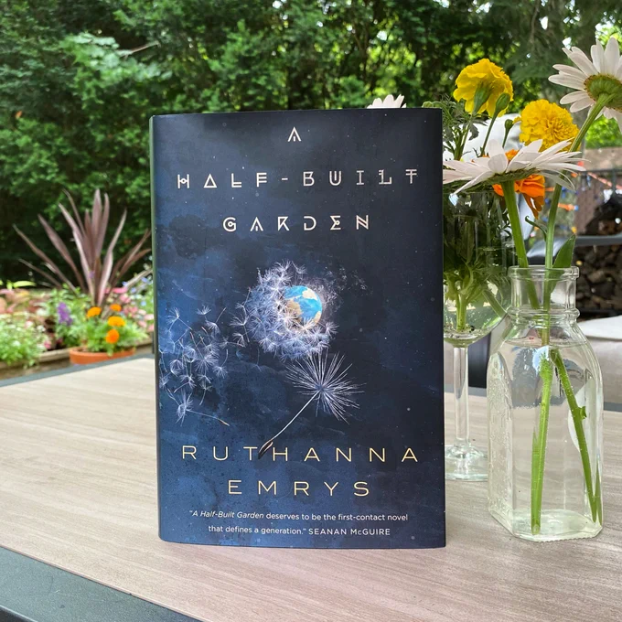 A copy of A Half-Built Garden by Ruthanna Emrys standing on top of a table next to a vase of flowers and with a garden in the background.