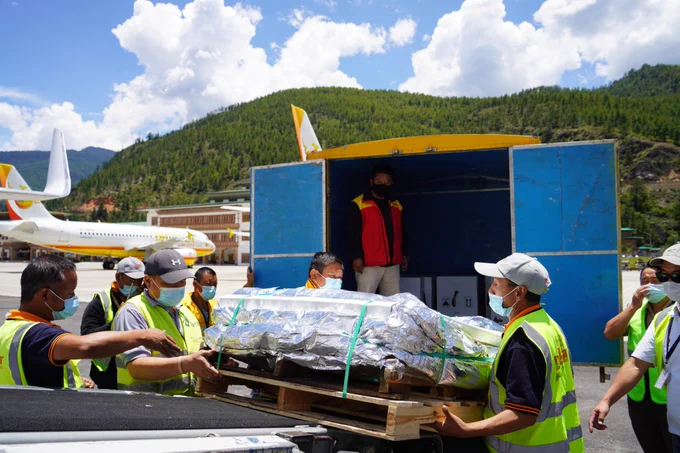 Airport crew members unload a pallet of insulated material from a cargo aircraft, with lush mountains in the background.