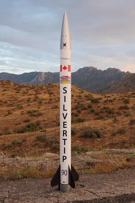 The team's rocket, wrapped in white vinyl, is standing upright in front of a landscape in New Mexico. It's name, Silvertip, is printed on it vertically, along with the flag of Canada and of British Columbia.