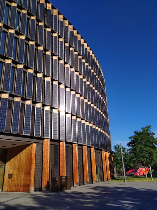 A modern administrative building with a curved façade. It is covered in thin solar panels and wood.