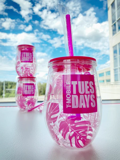 Clear cup decorated with vibrant magenta-colored palm leaves. The lid and straw are also magenta, branded with our magenta logo that says T-Mobile Tuesdays. One cup is in the foreground as the focus with a straw inserted, while two cups are stacked in the background. Behind the cups is a beautiful blue summer sky with fluffy white clouds.