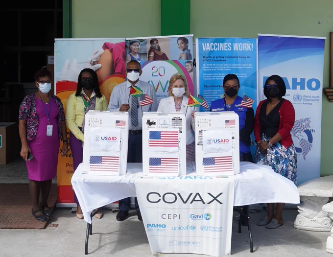 Five people stand behind a table, holding small American and Guyanese flags. On the table are boxes of COVID-19 vaccines labeled with the American flag and the words COVAX and USAID. Behind the people stand tall banners promoting vaccines.