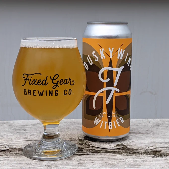 A glass of yellow beer sits on a wooden table next to a silver can with an orange label that has a brown butterfly on it. (Photo courtesy of Fixed Gear Brewing Co.)