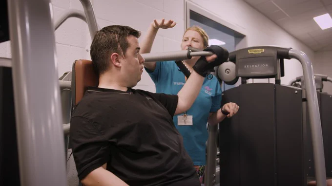 female personal trainer with CP, supporting a man on a gym machine.