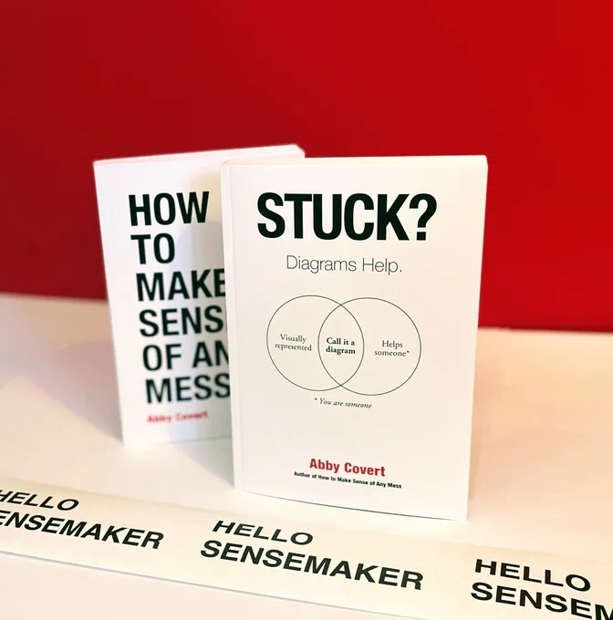 Two books, both by Abby Covert sit on a white surface with a red background. The books are titled "How to Make Sense of Any Mess" and "STUCK? Diagrams Help." There is also a section of packing tape shown that says "Hello Sensemaker".