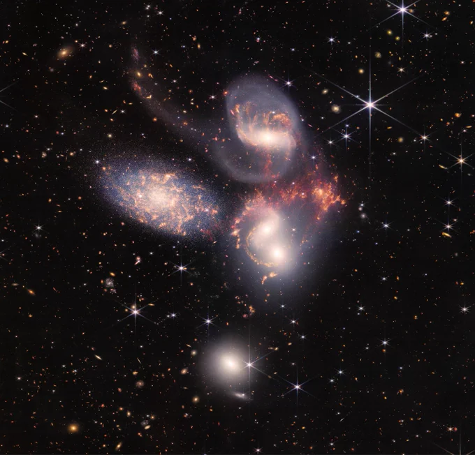 A group of five galaxies that appear close to each other in the sky: two in the middle, one toward the top, one to the upper left, and one toward the bottom. Four of the five appear to be touching. One is somewhat separated. In the image, the galaxies are large relative to the hundreds of much smaller (more distant) galaxies in the background. All five galaxies have bright white cores. Each has a slightly different size, shape, structure, and coloring. Scattered across the image, in front of the galaxies are number of foreground stars with diffraction spikes: bright white points, each with eight bright lines radiating out from the center.