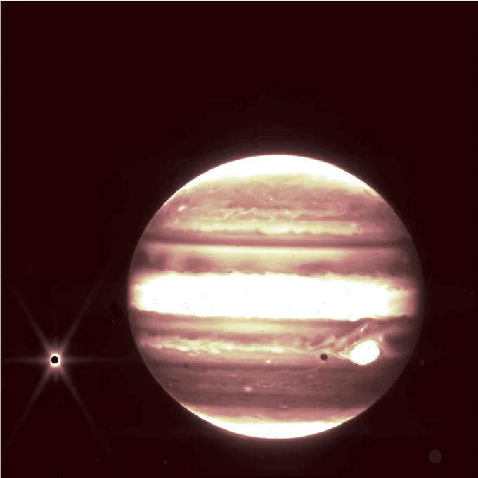 Jupiter dominates the frame, appearing to glow with bands of bright white, light yellow, and darker, brownish oranges. The stripes circle the planet, with one especially thick bright band across the planet’s center. A spot of glowing bright white interrupts the darker brown band about a third from the bottom of the planet. To the left of Jupiter, Europa appears as a tiny, black circle with a bright starburst erupting from its edges. The background of the image is pure black.