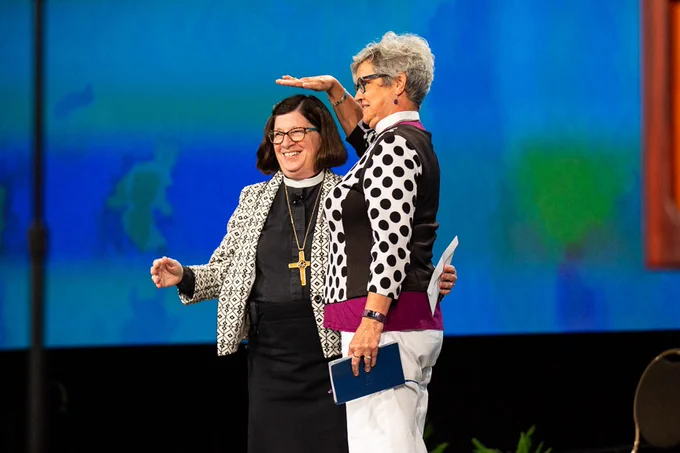 Evangelical Lutheran Church in Canada's National Bishop, Susan Johnson (right, white shirt with polka dots, clerical collar, white pants, and gray hair and glasses), playfully teases ELCA Presiding Bishop, Elizabeth Eaton (left, black shirt and skirt, white and black coat, clerical collar, gold cross, black hair and glasses) by holding her hand over Bishop Eaton's head. Both women are smiling as they laugh. 

A large blue screen with a couple splotches of green are behind the women. Photo was taken at ELCA Churchwide Assembly 2019.