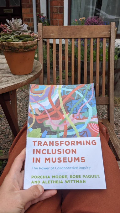 The cover of Transforming Inclusion in Museums: The Power of Collaborative Inquiry by Porchia Moore, Rose Paquet, and Aletheia Wittman, sitting on my lap in the garden with a brick terrace home with flower window boxes in the background. In the middle ground are patio furniture including a table with a potted succulent on it.