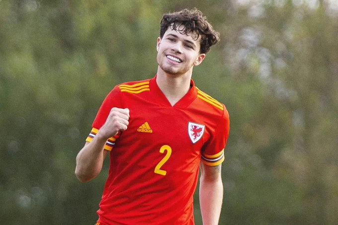 Neco Williams of Cymru is punching the air in a celebratory gesture following his goal during a UEFA Under-19 Championship Qualifying match against Russia in November 2019.

He is dressed in the red of Cymru, with yellow detailing on his shirt sleeves and shoulders. There is a large yellow 2 on his chest, along with the dragon crest of Cymru over his left pectoral muscle. 

Jogging towards the camera, Neco is smiling widely with embarrassingly perfect teeth.