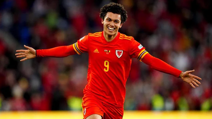 Brennan Johnson of Cymru celebrating after netting his first international goal four minutes from time, earning his country a dramatic 1-1 draw with Belgium in the Nations League competition.

He is dressed in the red of Cymru, with yellow detailing on his shirt sleeves and shoulders. There is a large yellow 9 on his chest, along with the dragon crest of Cymru over his left pectoral muscle. 

Running towards the camera, Brennan is smiling broadly with his arms held wide, palms open. 

He has a spectacular hairline.