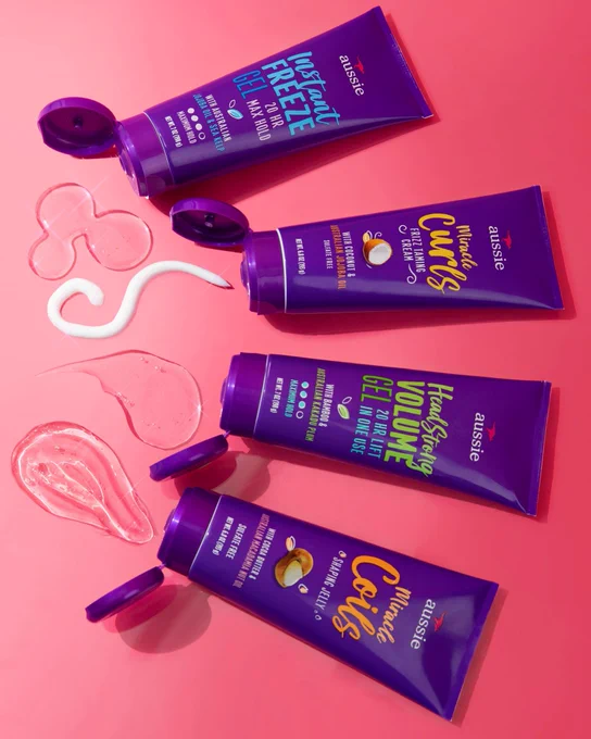 Aussie Instant Freeze Gel, Headstrong Volume Gel, Miracle Curls Frizz Taming Cream and Miracle Coils Shaping Jelly with product coming out of container on pink background.
