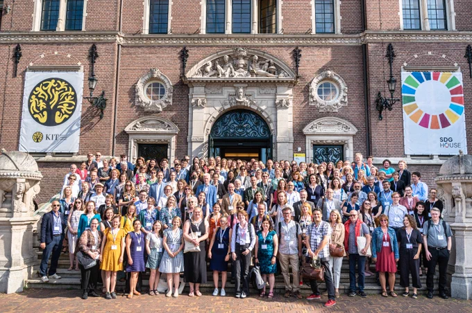 A photograph of a large group of people standing on the steps in front of Amsterdam's Royal Tropical Institute. The people stand in rows, wearing a variety of colours, looking happy. This group comprises many of the attendees of the SHARP 2022 conference. Two banners hand beside the stairs; the left banner featuring the Tropical Institute's logo, and the right banner featuring a colourful wheel with 'SDG House' written below.