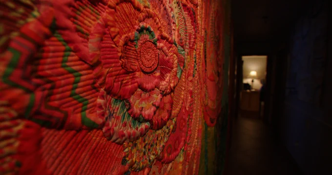 A rose patterned quilt hangs on the wall in the foreground of a hallway that falls into shadow. In the room at the end of a hall a woman sits by a desk lamp. 