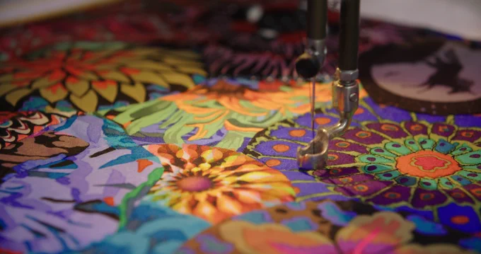Close up of a sewing machine knitting needle poised above a vibrantly patterned floral quilt. 